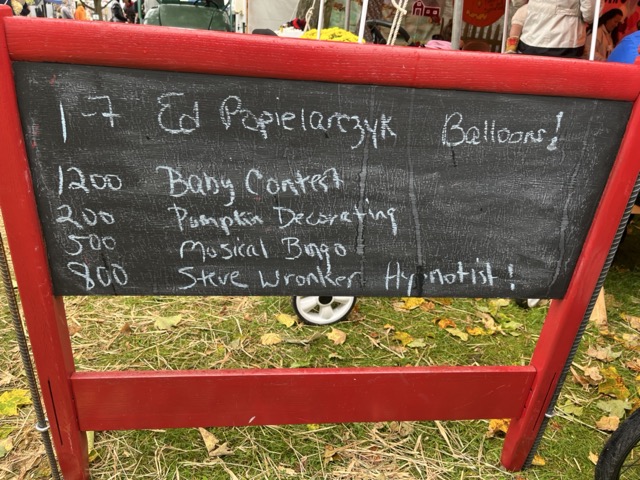 A hand-drawn sign in a folksy-fair environment that reads: 12:00 Baby contest, 2:00 Pumpkin decorating, 5:00 Musical bingo, 5:00 Steve Wronker hypnotist!